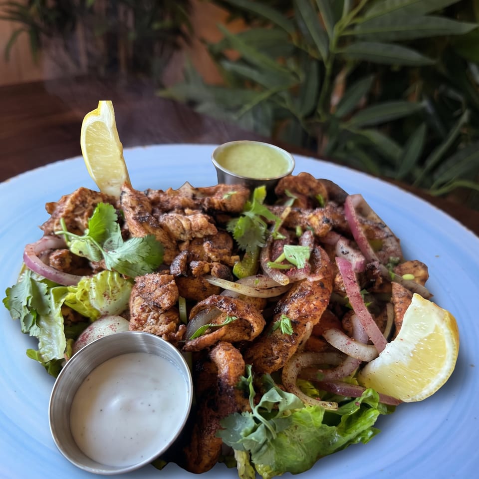 Grilled spiced meat pieces served with lettuce, cilantro, onions, lime wedges, and creamy dipping sauces on a blue plate.
