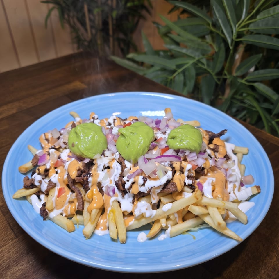 Loaded fries topped with avocado, chopped onions, grilled meat, and drizzled white and orange sauces on a blue plate.