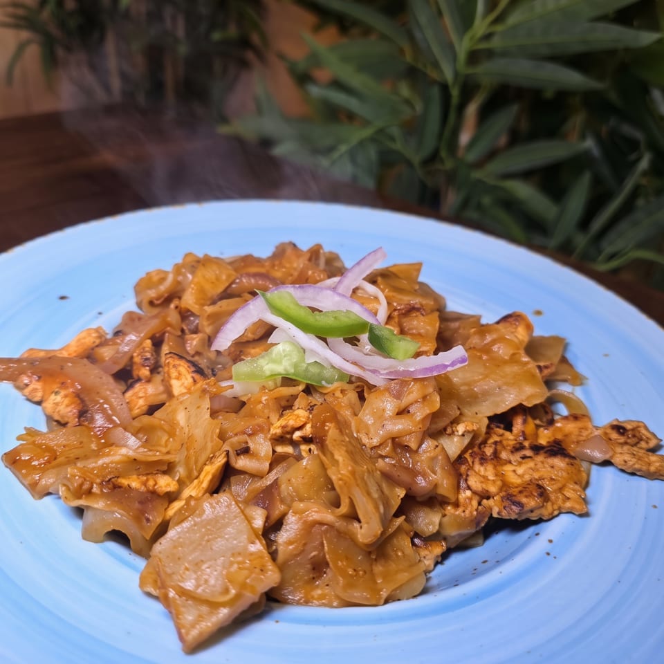 Creamy orange pasta with grilled chicken, red onion, and green pepper, served on a blue plate surrounded by plants.